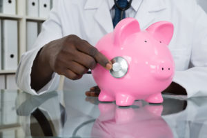 Close-up Of African Male Doctor Examining Piggybank With Stethoscope At Desk